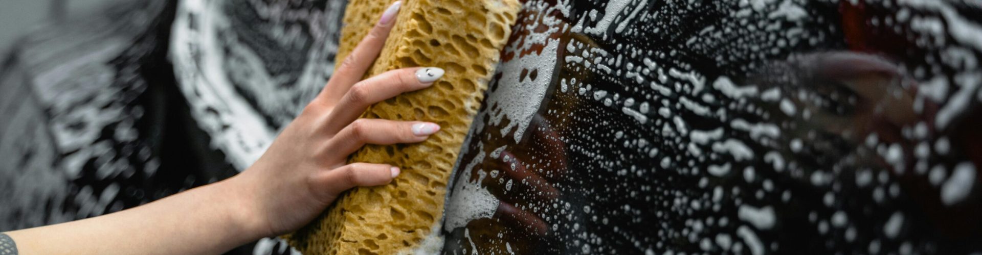 A hand scrubs a car window with a sponge, covered in soap and foam, at a car wash.